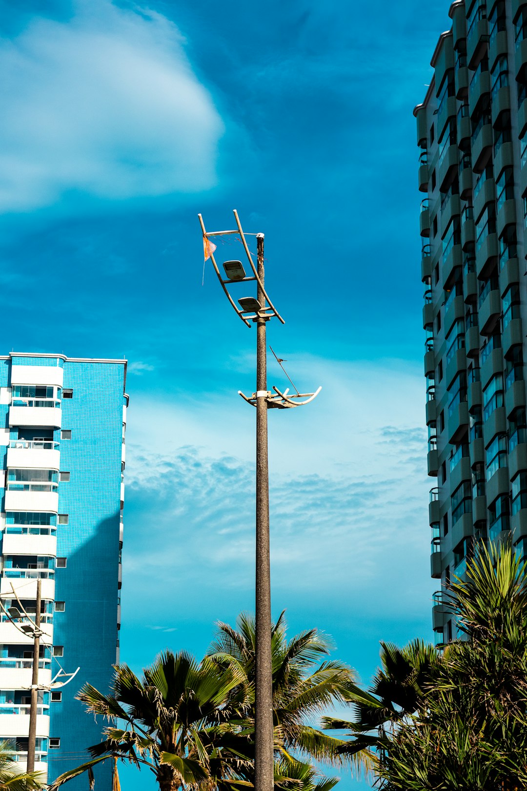 service-03 A tall, weathered lamppost stands between two modern apartment buildings under a bright blue sky with scattered clouds. Palm trees frame the bottom of the image, adding a tropical feel to the urban landscape.